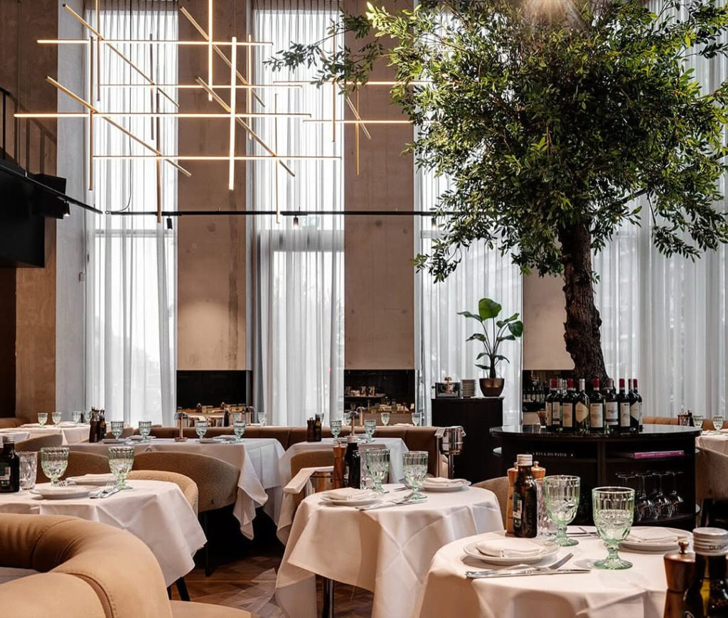 Doğal ışıkla aydınlanan yüksek tavanlı, modern bir İtalyan restoranının içinde, beyaz örtülü masalar, zarif açık kahverengi sandalyeler ve mekanın ortasında yükselen yemyeşil bir ağaç, Amsterdam seyahatinizde sizi bekleyen keyifli bir gastronomik durağı gösteriyor.