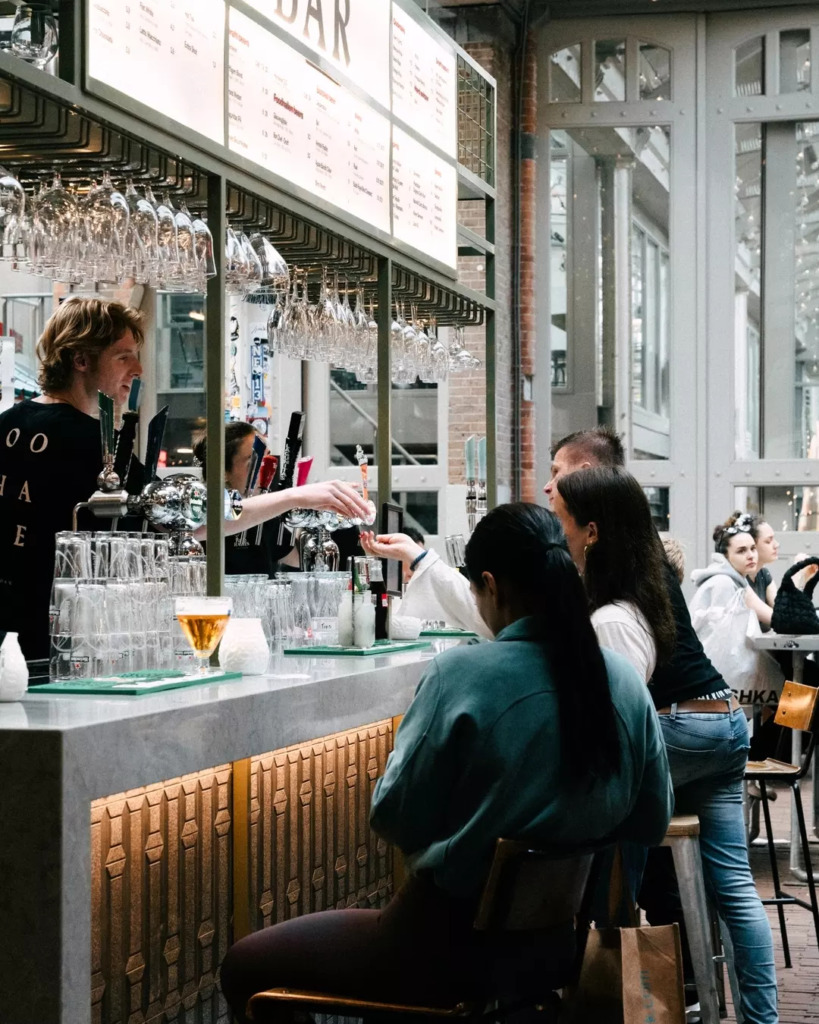 Amsterdam seyahati sırasında Foodhallen'in modern ve endüstriyel atmosferindeki hareketli bir barda, sarışın bir barmen müşterilere içki servisi yaparken, tavandan sarkan sayısız kadehin ve sohbetlerin eşlik ettiği keyifli bir gastronomik deneyim anı yansıtılıyor.