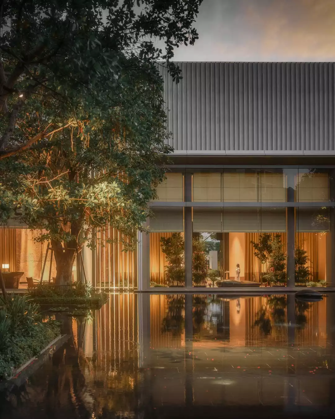 Yemyeşil ağaçların ve sıcak sarı ışıklarla aydınlatılmış geniş cam cephelerin dingin bir su havuzuna yansıdığı, zarif modern mimarisiyle öne çıkan Four Seasons Bangkok otelinin huzurlu bir dış görünümü.