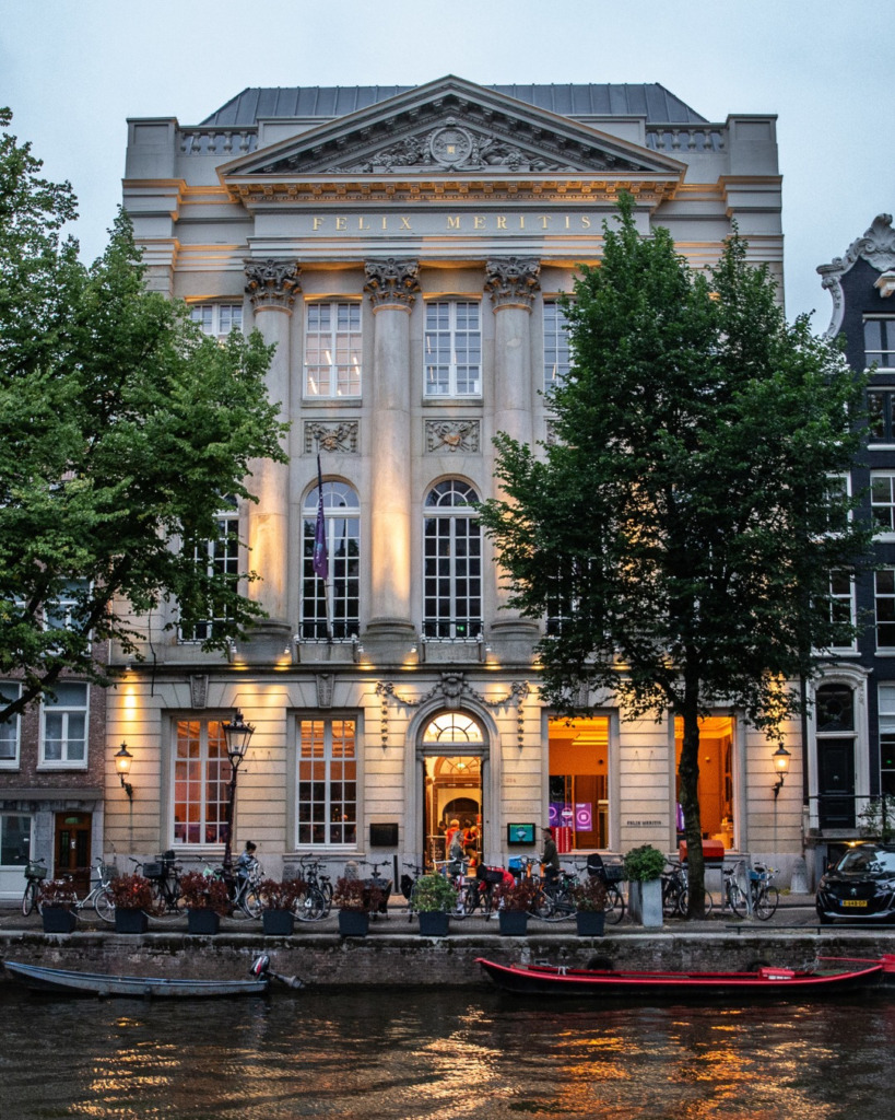 Akşamüstü, Amsterdam'ın tarihi kanallarından birinin kenarında, FELIX MERITIS yazılı, ışıklarla aydınlatılmış zarif neoklasik bina, önünde sıralanmış bisikletler ve suya yansıyan parıltılarla, şehrin sanat dolu ve büyülü atmosferini kusursuzca yansıtıyor.