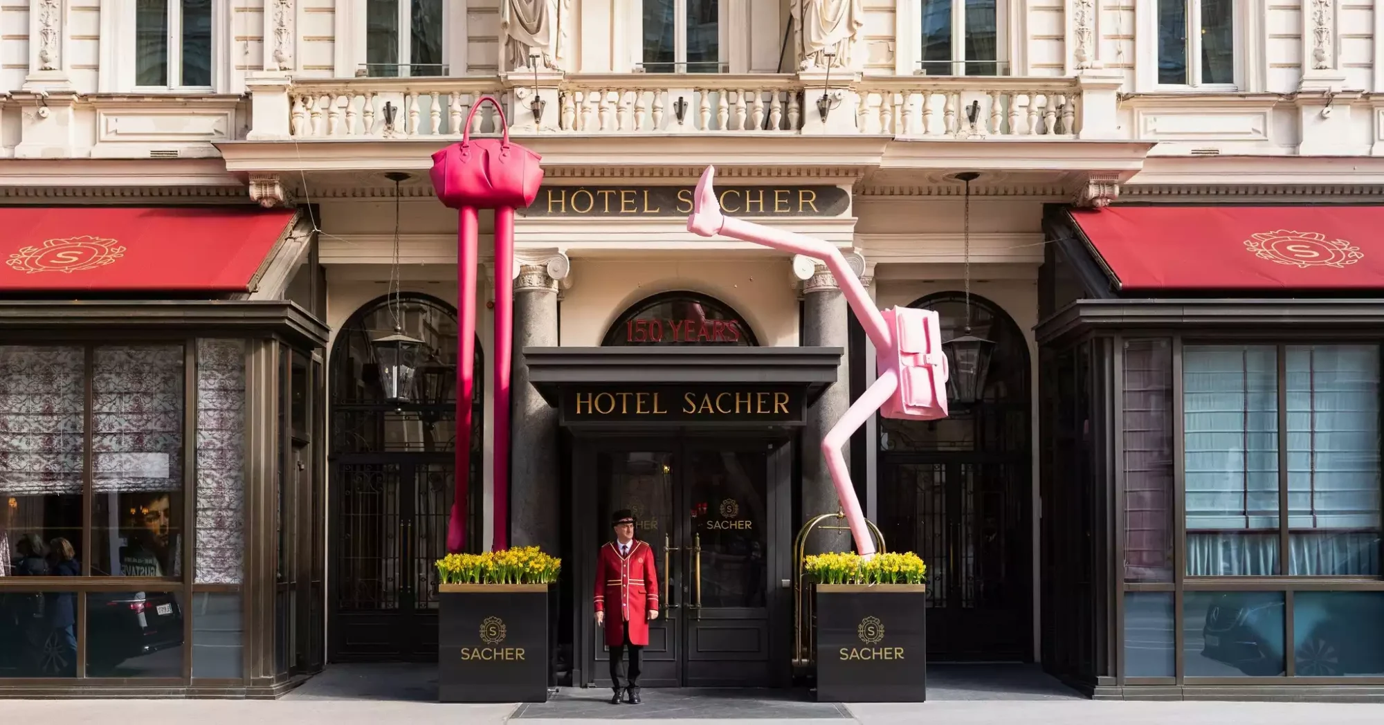 Viyana'daki lüks Hotel Sacher'in tarihi girişinde, bej renkli gösterişli cephesi ve kırmızı tenteleri önünde duran kapıcının iki yanında, pembe bir çanta ve sırt çantası detaylarıyla dikkat çeken, uzun ve sıra dışı sanat enstalasyonları otelin 150 yıllık köklü geçmişini modern bir dokunuşla yansıtıyor.
