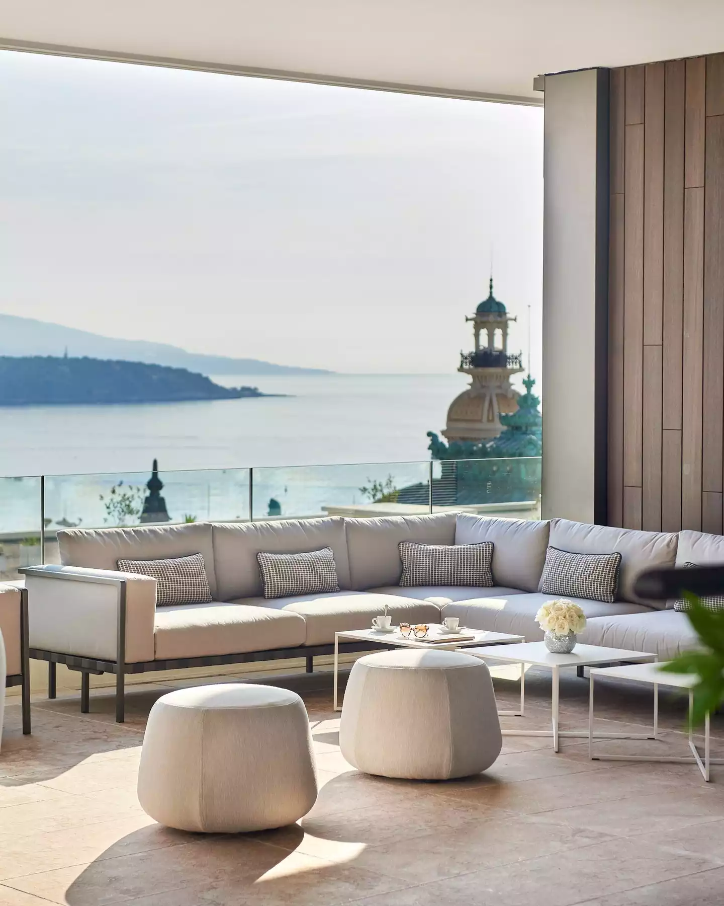 Güneşli bir günde, Hotel de Paris'in geniş terasında, Monte Carlo'nun masmavi denizine ve ikonik yeşil kubbeli binasına bakan lüks bir dinlenme köşesi; konforlu gri köşe koltuk takımı, beyaz puflar, üzerlerinde kahve fincanları ve beyaz güller olan sehpalar ile donatılmıştır.