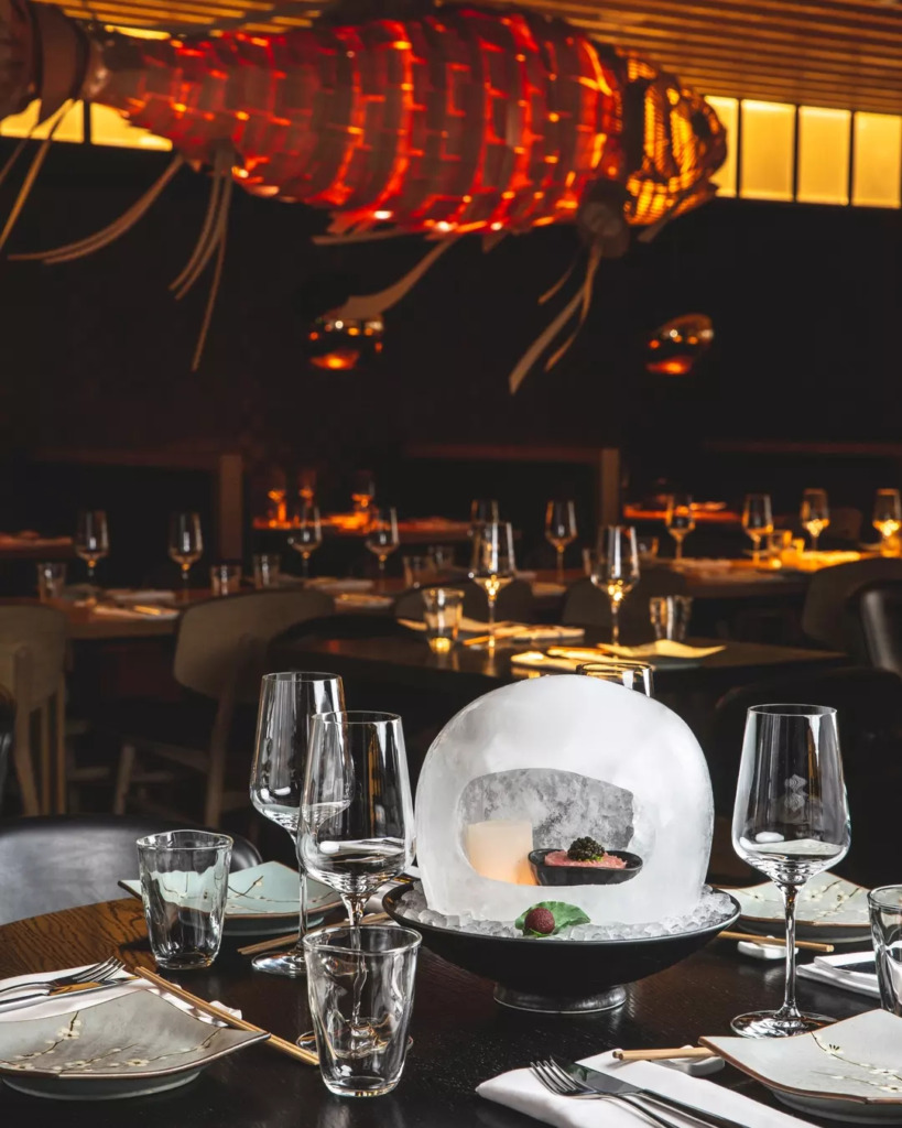 Amsterdam'ın De Pijp bölgesindeki Izakaya restoranında, loş ışıklar altında, Japon ve Latin Amerika mutfağını harmanlayan sarıkuyruk balığı saşimisinin havyar ve buz kubbe içinde sunulduğu şık bir masa düzeni ve tavanda asılı kırmızı balık figürlü avize, şehrin canlı gece hayatı ve gastronomi duraklarından birini yansıtıyor.