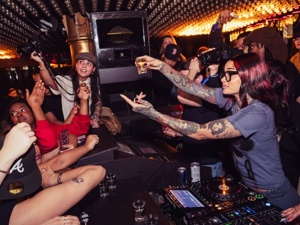 Yüksek tavanlı, ışıklarla süslü bir kulüpte, kırmızı saçlı ve dövmeli bir DJ, Amsterdam Seyahat Rehberi'nde bahsedilen şehrin enerjik gece hayatı deneyimlerinden birini yansıtır şekilde, elinde bir shot bardağıyla coşkulu kalabalığa doğru işaret ederek müzik çalıyor ve etrafındaki herkesin keyifli anlar yaşadığı bu ortamda eğlence doruklara ulaşıyor.