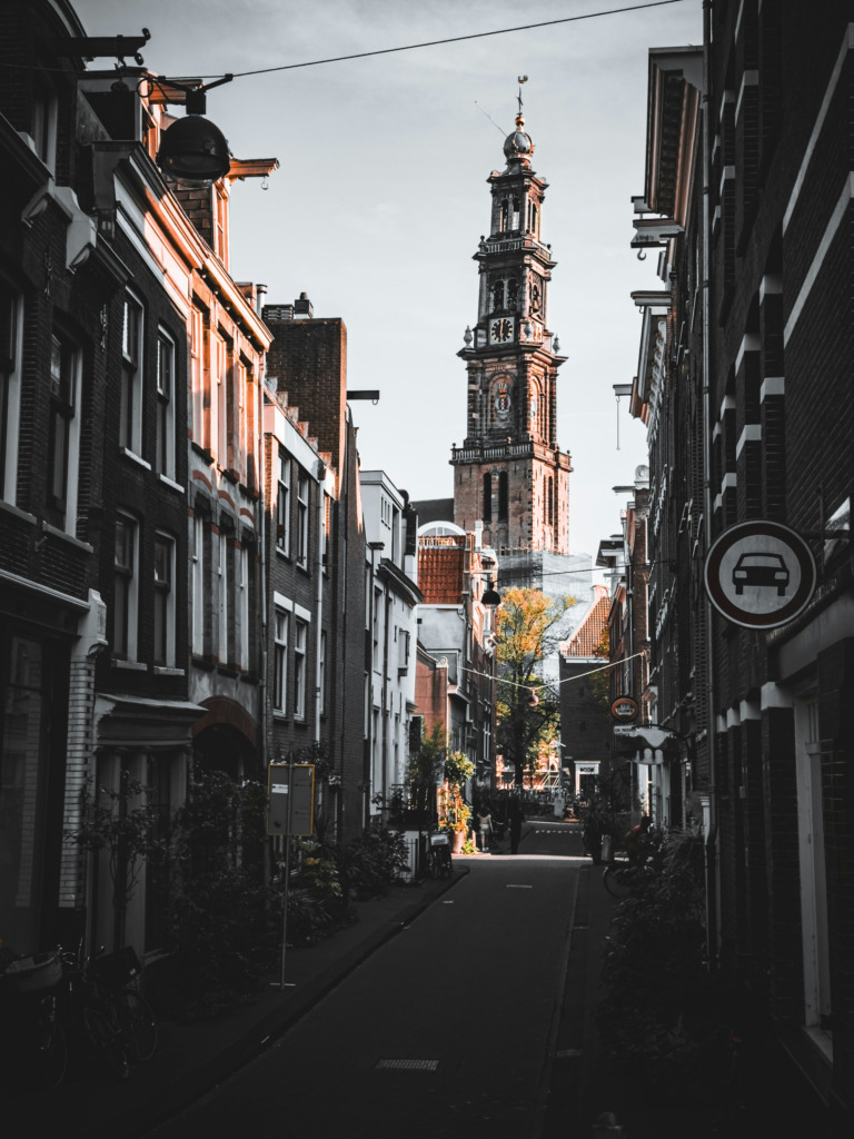 Amsterdam Seyahat Rehberi'nde bahsi geçen Jordaan Mahallesi'nin, gün ışığıyla parlayan tuğla cepheli dar sokaklarından uzakta yükselen, şehrin tarihi dokusunu simgeleyen oymalı bir kilise kulesi görünüyor.