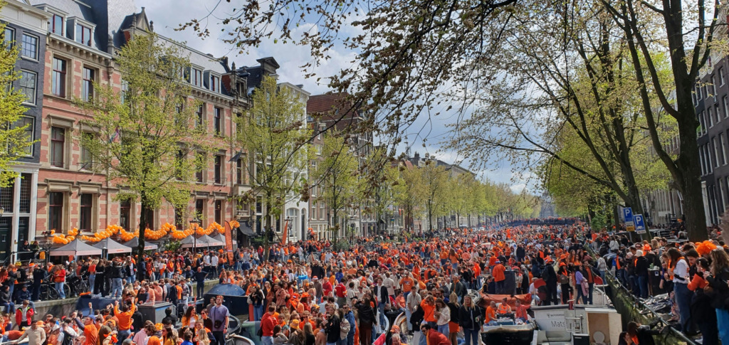 Baharın taze yeşillikleriyle süslenmiş ağaçların gölgelediği Amsterdam kanallarından birinde, turuncu kıyafetler içindeki binlerce insanın Koningsdag kutlaması için toplanıp teknelerle ve sokaklarda neşeyle eğlendiği, şehrin enerjik ve rengarenk atmosferini yansıtan kalabalık bir görünüm.