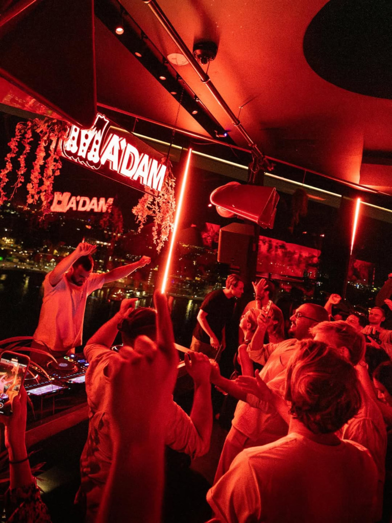 M'ADAM kulübünde, DJ performansı eşliğinde kırmızı ışıklarla aydınlanmış bir ortamda eğlenen kalabalık, pencereden görünen Amsterdam'ın ışıltılı gece manzarası ve kanallar üzerinde yansıyan ışıklarla şehrin hareketli gece hayatını canlı bir şekilde yansıtıyor.
