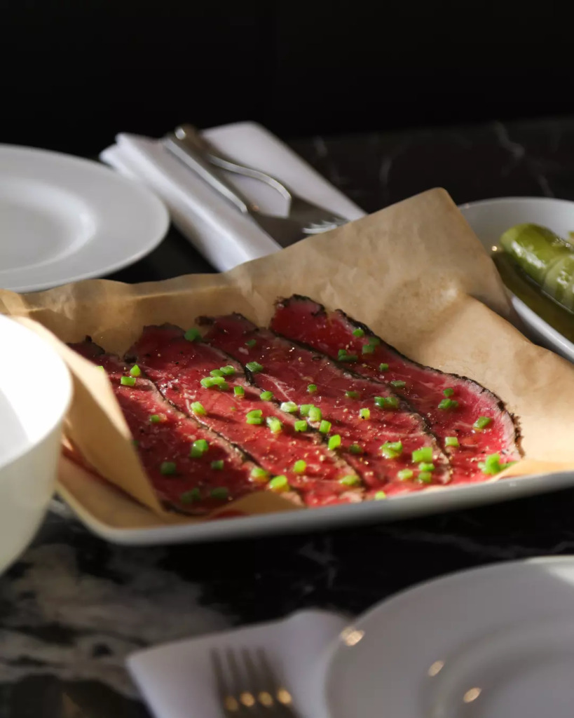 Mr Porter restoranında koyu renkli mermer bir masada, parşömen kağıdı üzerinde sunulan, doğranmış yeşil soğanlarla zenginleştirilmiş ince dilimlenmiş wagyu tartar, Amsterdam'ın zarif akşam yemekleri için önerilen seçkin lezzetlerinden biri.
