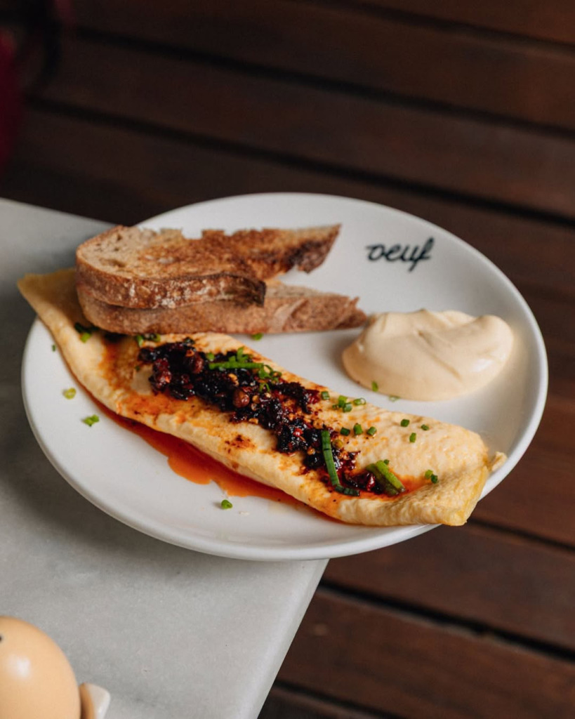 Beyaz bir tabakta, üzerinde baharatlı ve kıtır sos, taze soğan ile zenginleştirilmiş bir omlet, yanında kızarmış ekmek dilimleri ve kremalı bir sos ile sunulan bu kahvaltı tabağı, Amsterdam seyahat rehberi kapsamında Oeuf’un yaratıcı ve doyurucu bir seçeneğini gösteriyor.