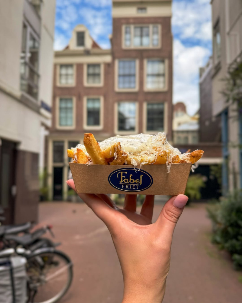 Bir el, Amsterdam'ın karakteristik tuğla binaları ve dar sokaklarının hafifçe bulanık göründüğü arka planda, Fabel Friet logosu taşıyan karton kutuda trüflü mayonez ve rendelenmiş peynirle zenginleştirilmiş, şehrin sevilen sokak lezzetlerinden çıtır patates kızartması tutuyor.