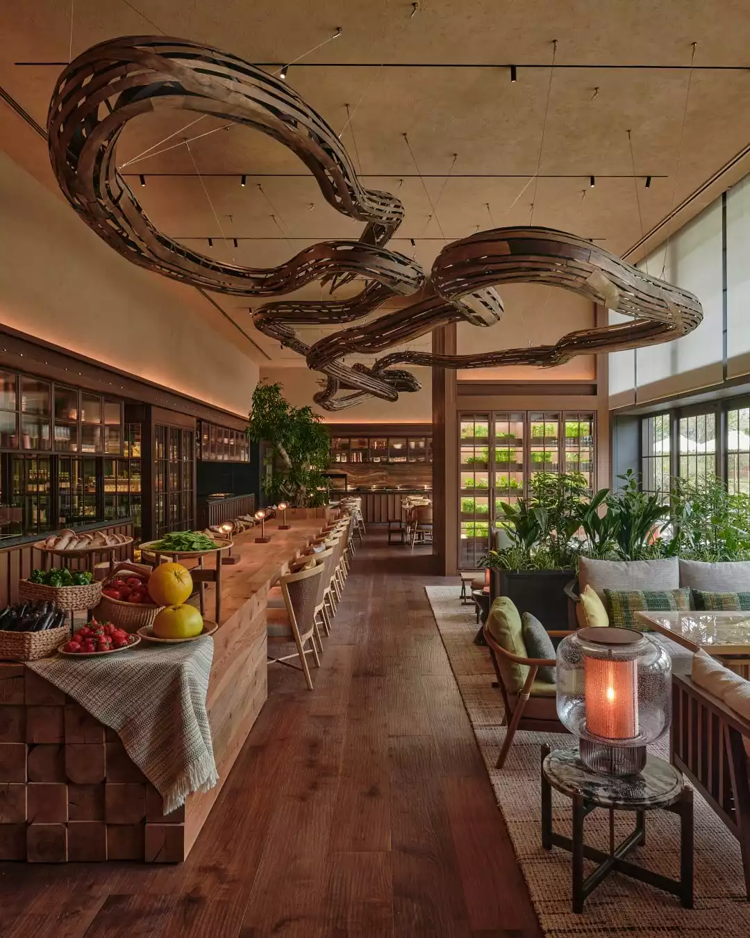 Patina Osaka restoranının geniş iç mekanında, taze ürünlerle donatılmış uzun bir açık büfe tezgahı, yemyeşil bitkilerle çevrili rahat bir oturma alanı ve tavanı süsleyen dalgalı, büyük ahşap aydınlatma armatürleri modern ve doğal bir atmosfer yaratıyor.