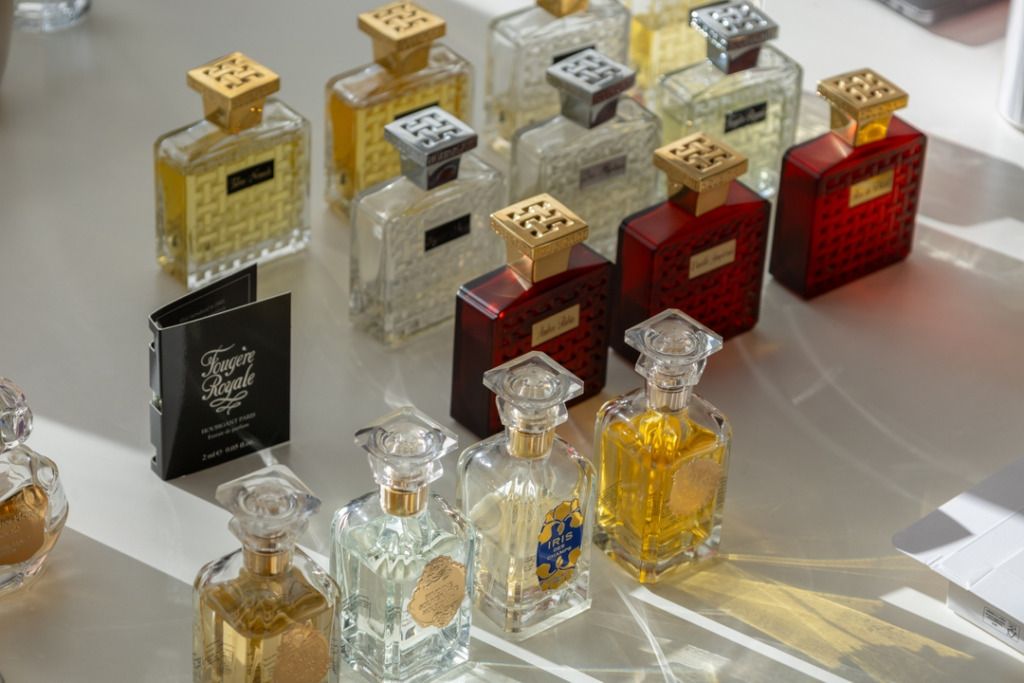Perfume Lounge'un zarif atmosferini yansıtan bu görselde, güneş ışığı alan beyaz bir yüzeyde, altın ve gümüş kapaklı, farklı renklerdeki lüks niş parfüm şişeleri şık bir biçimde sıralanmış, Amsterdam seyahat rehberi için özel bir alışveriş önerisini gözler önüne seriyor.