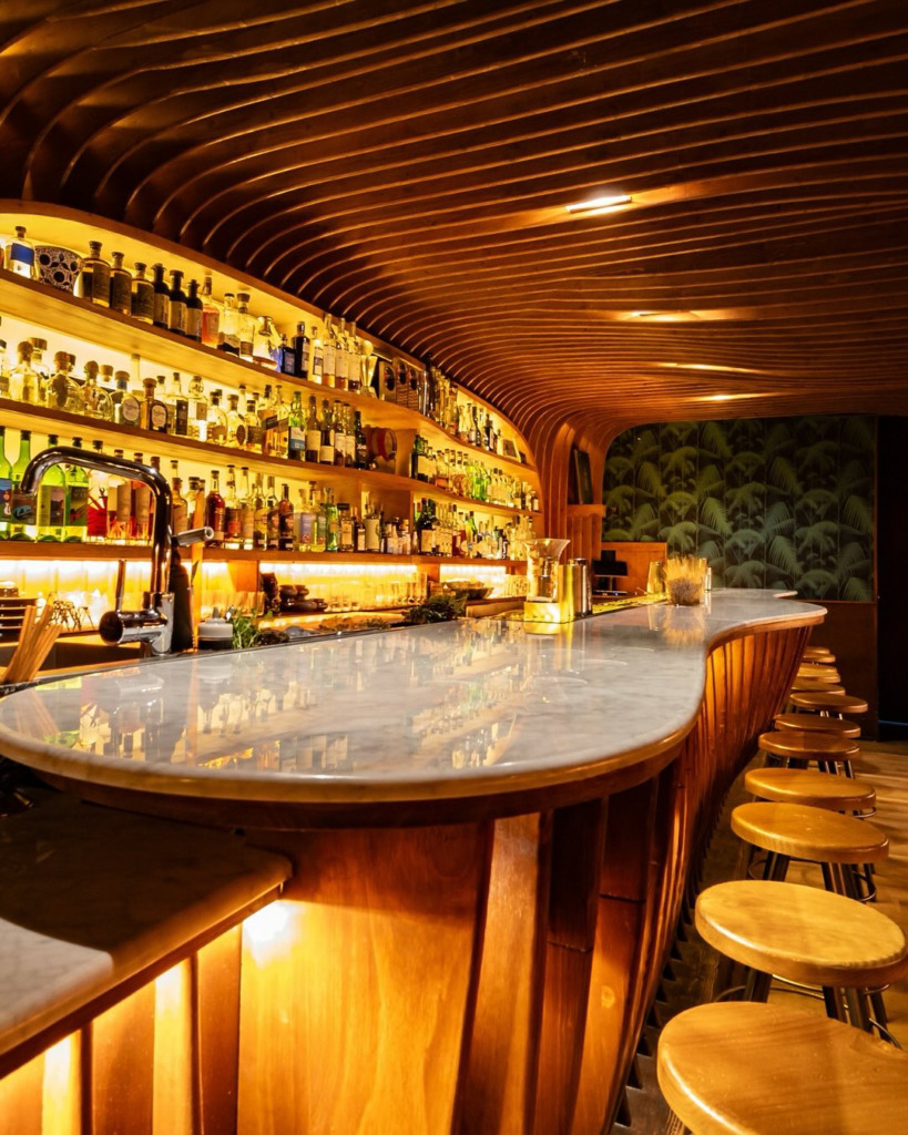 Amsterdam’daki Pulitzer Bar’ın Art deco zarafetiyle tasarlanmış ortamında, kavisli mermer bar tezgâhı, ahşap latalarla kaplı dalgalı tavanı, arkadan aydınlatılmış şişelerle dolu rafları ve ahşap bar tabureleri sıcak bir atmosfer sunuyor.