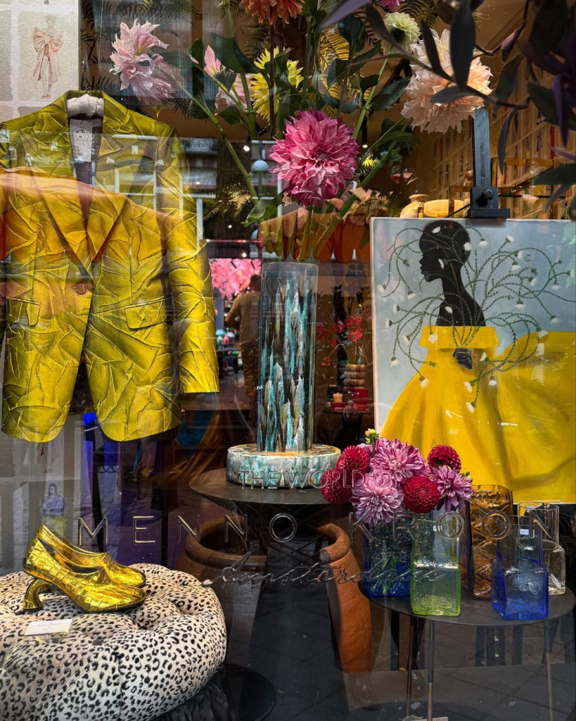 Amsterdam'ın lüks ve sanatsal ruhunu yansıtan bir butik vitrininde, kabartmalı parlak sarı bir ceket, leopar desenli puf üzerinde duran altın rengi topuklu ayakkabılar, sarı elbiseli bir kadın silüeti tablosu ve canlı çiçek aranjmanları modern Hollanda tasarımını gözler önüne seriyor.