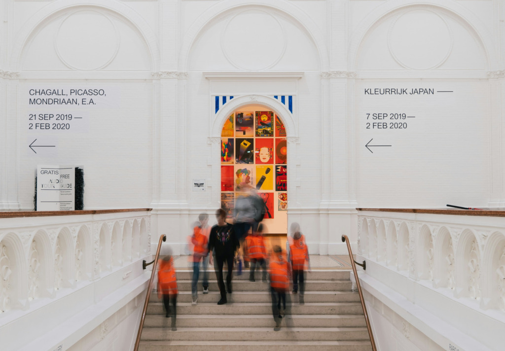 Amsterdam Seyahat Rehberi'nde öne çıkan modern sanatın merkezlerinden Stedelijk Müzesi'nin ışık dolu, kemerli beyaz bir salonunda, Chagall, Picasso ve Mondriaan gibi ustaların eserlerine ve renkli Japonya sergisine işaret eden tabelaların önünden, turuncu yelekli ziyaretçiler hareket bulanıklığıyla aşağı merdivenlerden inerken şehrin sanatla iç içe ruhunu yansıtıyor.
