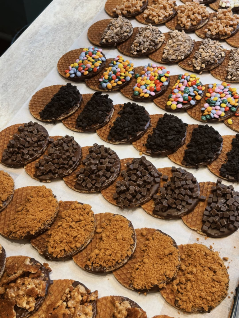 Amsterdam'ın özgün sokak lezzetlerinden stroopwafeller, görselde çikolataya batırılarak üzerine rengarenk şekerlemeler, bisküvi kırıntıları, minik çikolata küpleri ve karamelize edilmiş farklı parçacıklar serpilmiş haliyle iştah açıcı bir sunumla sergileniyor.