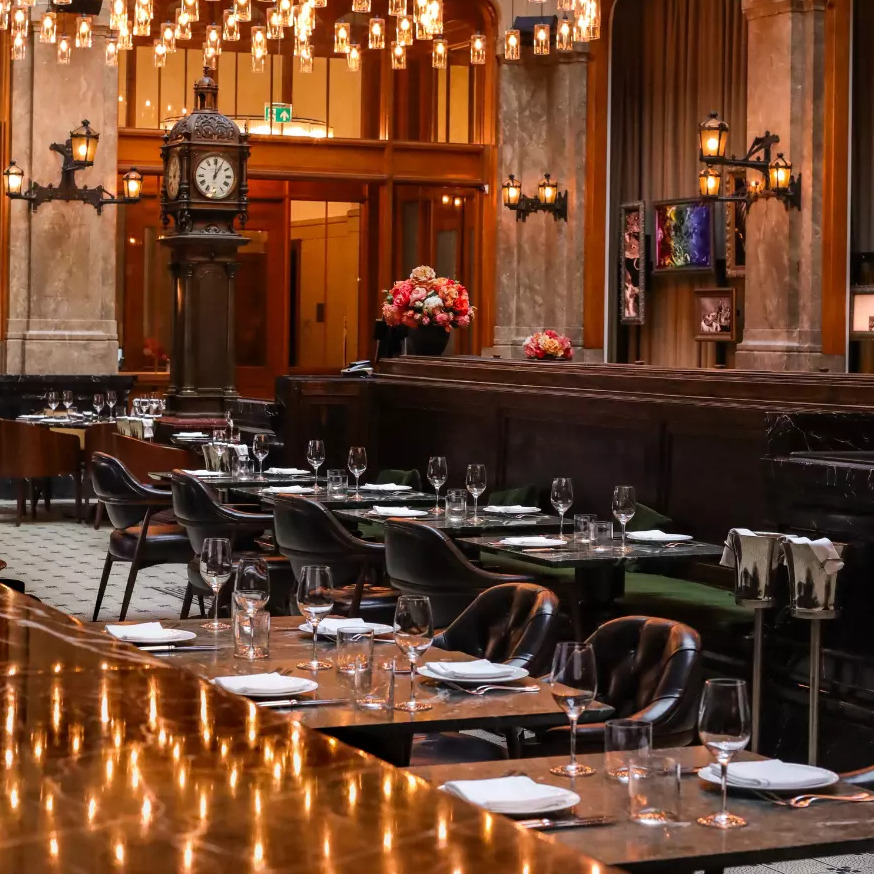Amsterdam'ın zengin Belle Époque döneminden kalma eski bir banka binasında yer alan, yüksek tavanlı, parıldayan avizeler ve zarif detaylarla bezeli The Duchess restoranının iç görünümü, şık masaları ve gösterişli duvar saatiyle misafirlerini unutulmaz bir deneyime davet ediyor.