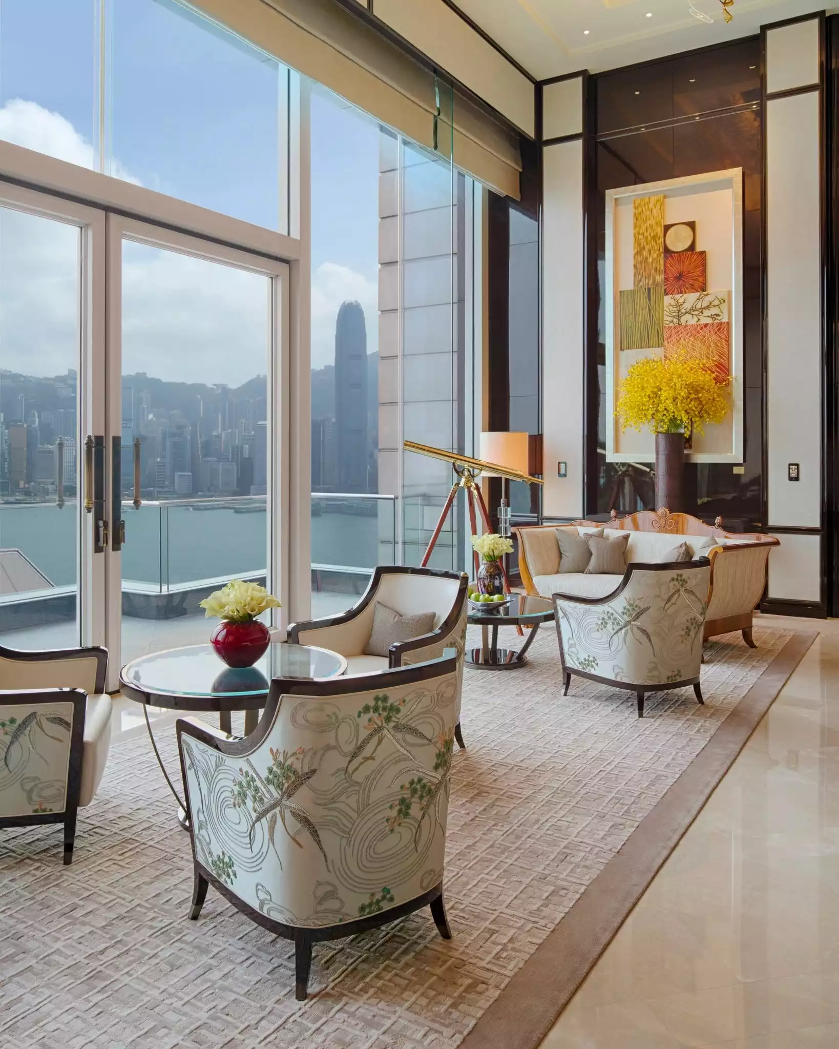 The Peninsula Hong Kong'daki lüks bir yaşam alanında, Hong Kong şehrinin ve limanının nefes kesen manzarasına sahip büyük pencereler önünde, kuş desenli zarif koltuklar, pirinç bir teleskop ve canlı çiçek aranjmanlarıyla donatılmış şık bir oturma düzeni sunulmaktadır.