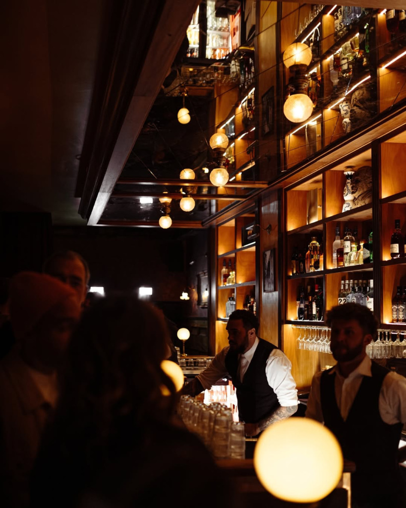 Ahşap raflarda parıldayan içki şişeleri ve yansımalı tavandan sarkan yuvarlak, sıcak ışıklarla aydınlanan, Amsterdam gece hayatının canlı duraklarından Tuf Tuf barında, barmenlerin özenle kokteyller hazırladığı, müşterilerin keyifli sohbetler eşliğinde vaktini geçirdiği samimi ve hareketli bir an.
