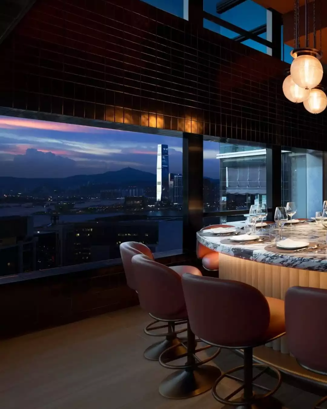 Upper House Hong Kong'un şık iç mekanında, mermer bir bar tezgahı ve konforlu deri sandalyeler, gün batımının büyüleyici mor ve turuncu tonlarıyla aydınlanan Hong Kong şehir silüeti ve uzak dağların nefes kesen panoramik manzarasına karşı konumlanmış.