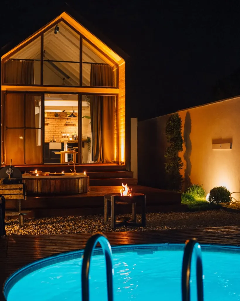 Gece karanlığında, geniş cam cephesinden ışıklar saçan modern ahşap bungalov, önündeki parlak mavi havuz, mumlarla aydınlatılmış sıcak küvet ve yanan ateş çukuruyla huzurlu ve davetkar bir dinlenme ortamı sunuyor.