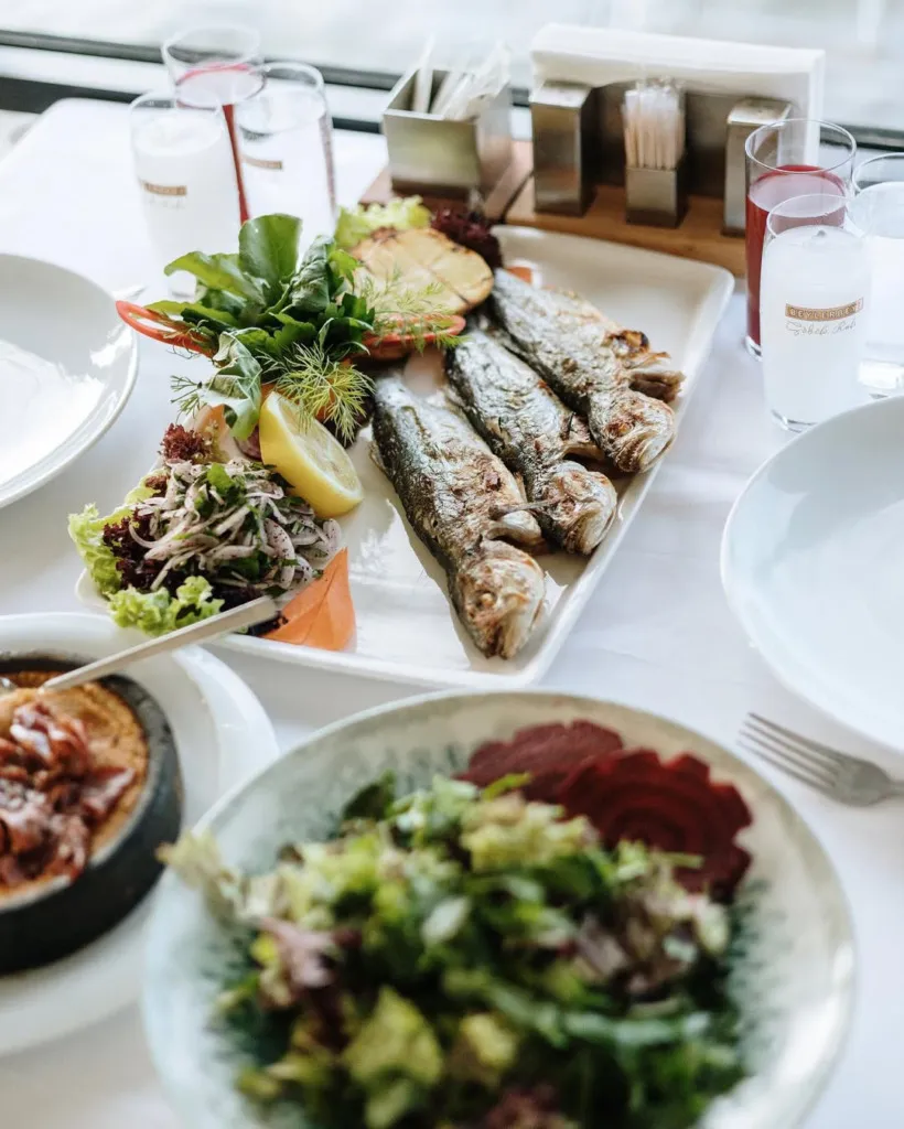 Sapanca Gölevi Resort'un restoranında beyaz masa örtüsü üzerinde dört adet ızgara balık, bol yeşillikli salatalar, taze limon dilimi ve çeşitli içeceklerle donatılmış, misafirlere keyifli bir ziyafet sunan iştah açıcı bir sofra.