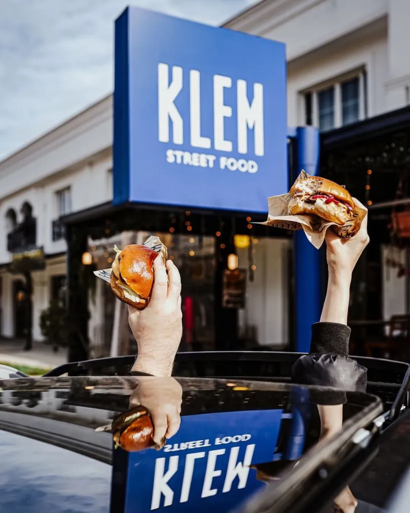 KLEM Street Food tabelasının önünde, bir arabanın sunroofundan uzanan iki el, pofuduk parlak ekmekli ve soslu KLEM burgerlerini havaya kaldırarak Sapanca'daki dinamik sokak yemeği kültürünü yansıtıyor.