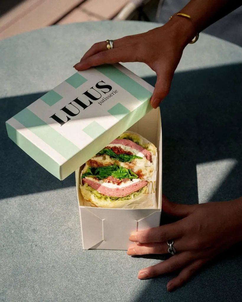Bir kişi, üzerinde LULUS patisserie logosu ve açık mint yeşili çizgiler bulunan beyaz bir kutuyu açarken, kutunun içinden pembe şarküteri dilimleri, beyaz peynir, kurutulmuş domates, yeşillikler ve pesto sosuyla zenginleştirilmiş iştah açıcı bir tuzlu pasta veya sandviç görünüyor.