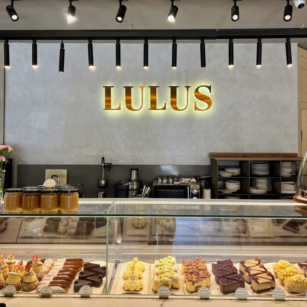 LULUS adlı modern pastanenin iç görünümünde, ışıklı altın harfli LULUS tabelasının altında, cam vitrin içerisinde iştah açıcı cheesecake'ler, eklerler ve çeşitli pastaların sergilendiği bir tezgah ve arkasında mutfak ekipmanları ile düzenli raflar görülüyor.