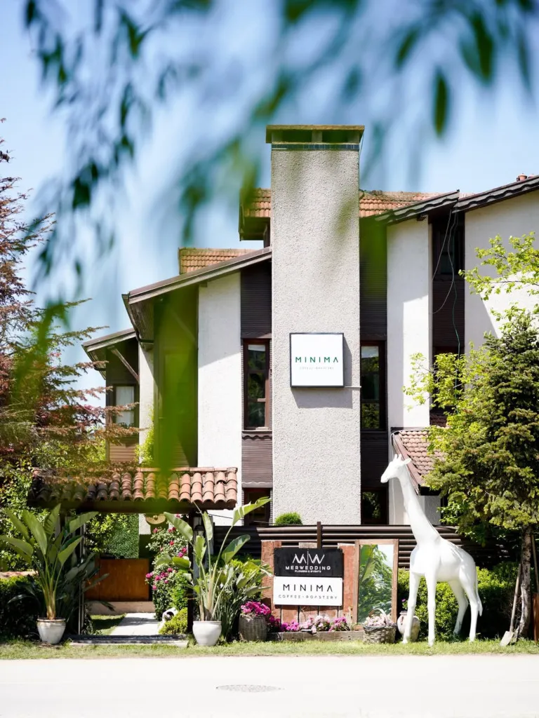 Minima Coffee Roastery'nin adının yazılı olduğu modern binanın önünde, Minima Coffee ve Merwedding tabelaları, uzun beyaz zürafa heykeli, canlı yeşil bitkiler ve kiremitli kemerli giriş, Minima Coffee Sapanca'nın davetkar dış cephesini ve özenli çevresini gözler önüne seriyor.