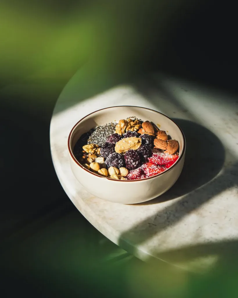 Dondurulmuş böğürtlenler, chia tohumları, cevizler, bademler, fındıklar, fıstık ezmesi ve çilek dilimleriyle dolu besleyici bir kase, Minima Coffee gibi bir mekanda mermer masada güneş ışığı altında, yeşil bitki detaylarıyla ferahlatıcı bir kahvaltı anı sunuyor.