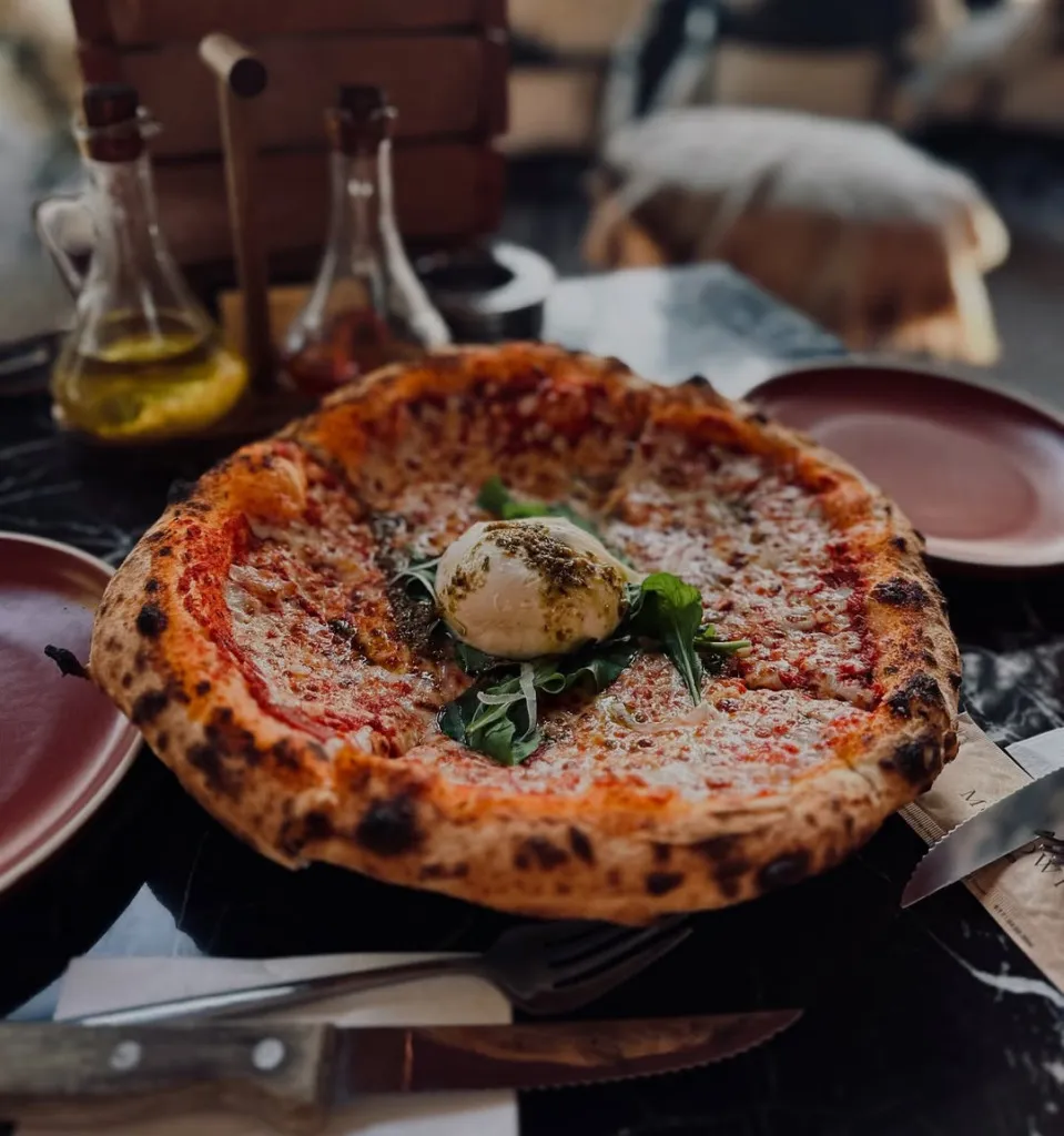 Sapanca'da bir masada duran, kenarları belirgin şekilde pişmiş hamurlu, bol mozzarella ve domates soslu, ortasında kremsi burrata peyniri, pesto sosu ve taze roka yapraklarıyla süslenmiş, iştah açıcı bir pizza.