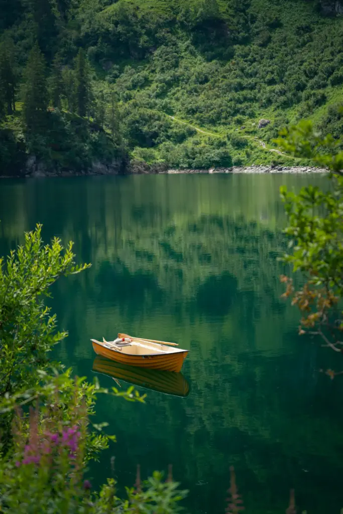 Yemyeşil tepelerin ve sık ormanların çevrelediği berrak, koyu yeşil bir gölün dingin sularında, kürekleri içinde sakince yüzen turuncu bir sandalın eşsiz yansımasıyla huzur dolu bir doğa gezi noktası.