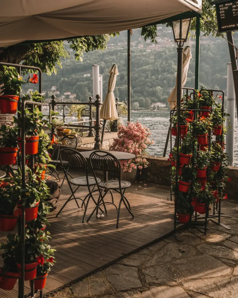 Sapanca gezi rehberi durağı olarak tercih edilebilecek, kırmızı saksılı çiçekler ve pembe açan bitkilerle süslü, göl kenarında ahşap zeminli bir açık hava kafe alanı, arka planda yeşil tepeler ve sakin su manzarasıyla huzurlu bir dinlenme sunuyor.