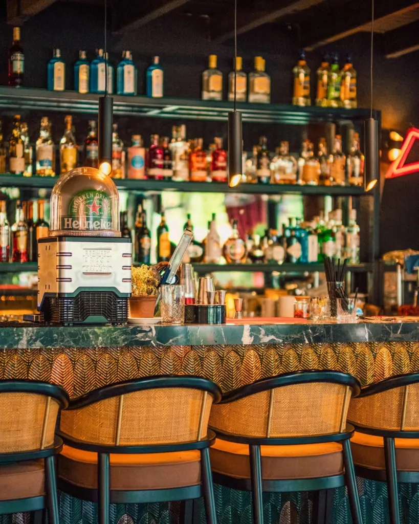 Koyu yeşil mermer bar tezgahında duran Heineken ışıklı tabelası, hasır tabureleri ve arkadaki bol çeşitli içki raflarıyla modern bir bara ait bu detaylı çekim, Sapanca Palmiye Homes gibi konut projelerinde sunulan sosyal alanların zenginliğini ve tarzını vurguluyor.