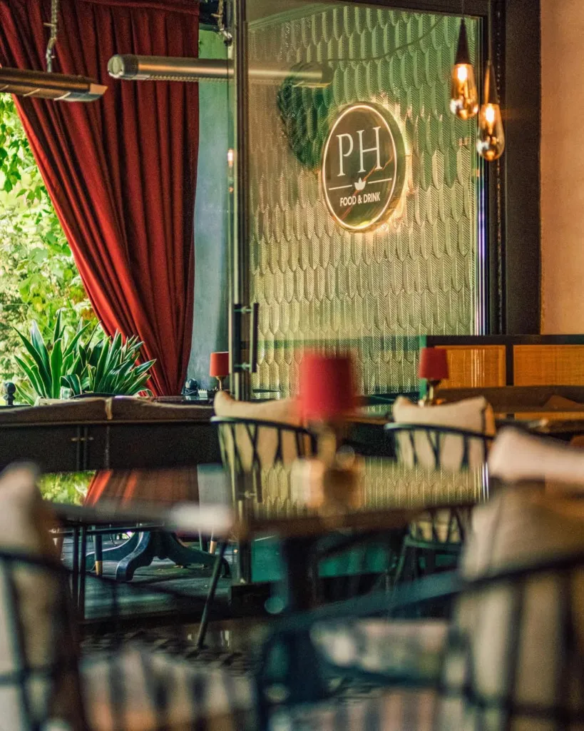Dışarıdaki yemyeşil doğayla çevrili Sapanca Palmiye Evleri bünyesindeki PH FOOD & DRINK restoranının, desenli cam kapısında ışıklı logosu yansıyan, kırmızı perdeler ve zarif oturma gruplarıyla dolu davetkar ve şık iç mekanı.