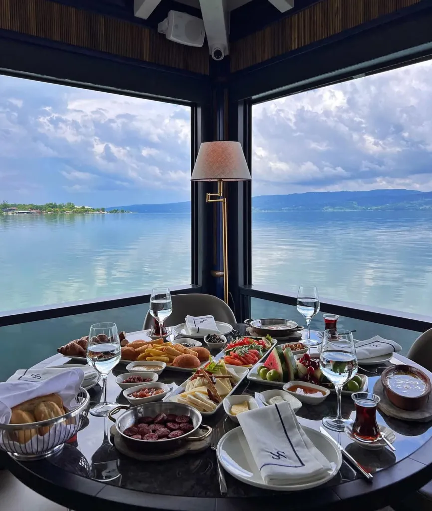 Sasa Restoran'ın Sapanca Gölü'ne nazır panoramik penceresinin önünde, sucuk tava, çeşitli peynirler, zeytinler, reçeller, ekmek çeşitleri, patates kızartması ve meyvelerle dolu zengin bir Türk kahvaltısı masası, bulutlu gökyüzü ve uzak dağlar eşliğinde göz alıcı bir manzara sunuyor.