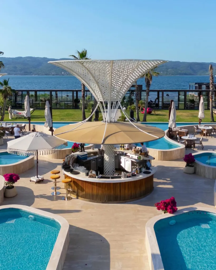 Sapanca'da göle nazır, lüks bir resort otelinde modern ve süslü beyaz bir üst yapıya sahip şık bir dış mekan barı, etrafında eğimli kenarlı turkuaz havuzlar, güneş şemsiyeleri, palmiye ağaçları ve oturma alanları ile ferahlatıcı bir yaz günü manzarası.