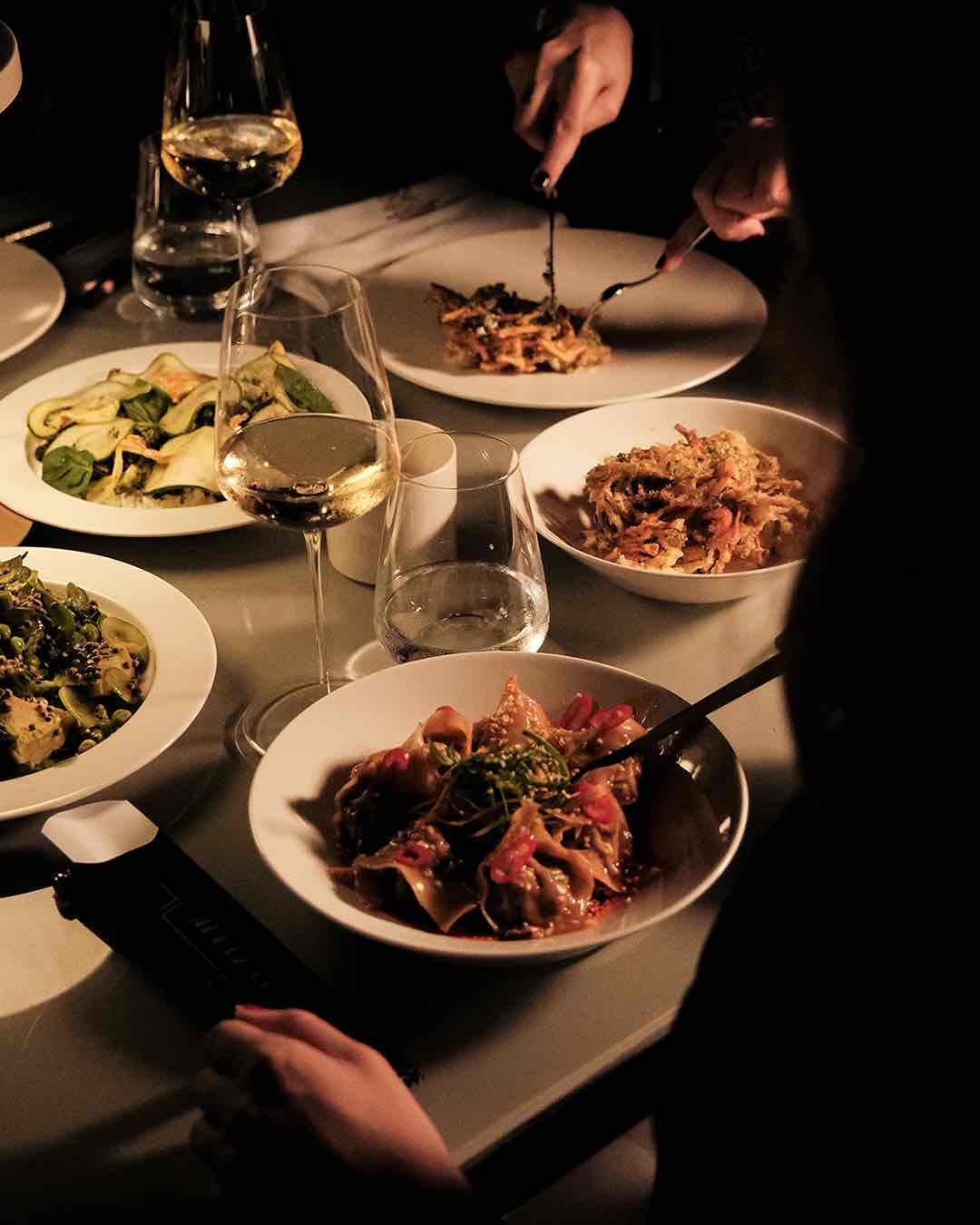 Loş ışıklandırılmış bir restoranda veya yemek masasında, beyaz tabaklarda sunulmuş çeşitli yemekler (makarna, soslu bir ana yemek ve salata gibi), şarap kadehleri ve su bardaklarıyla dolu samimi bir akşam yemeği anı; bir misafir yemeğini çatal bıçak kullanarak yiyor.