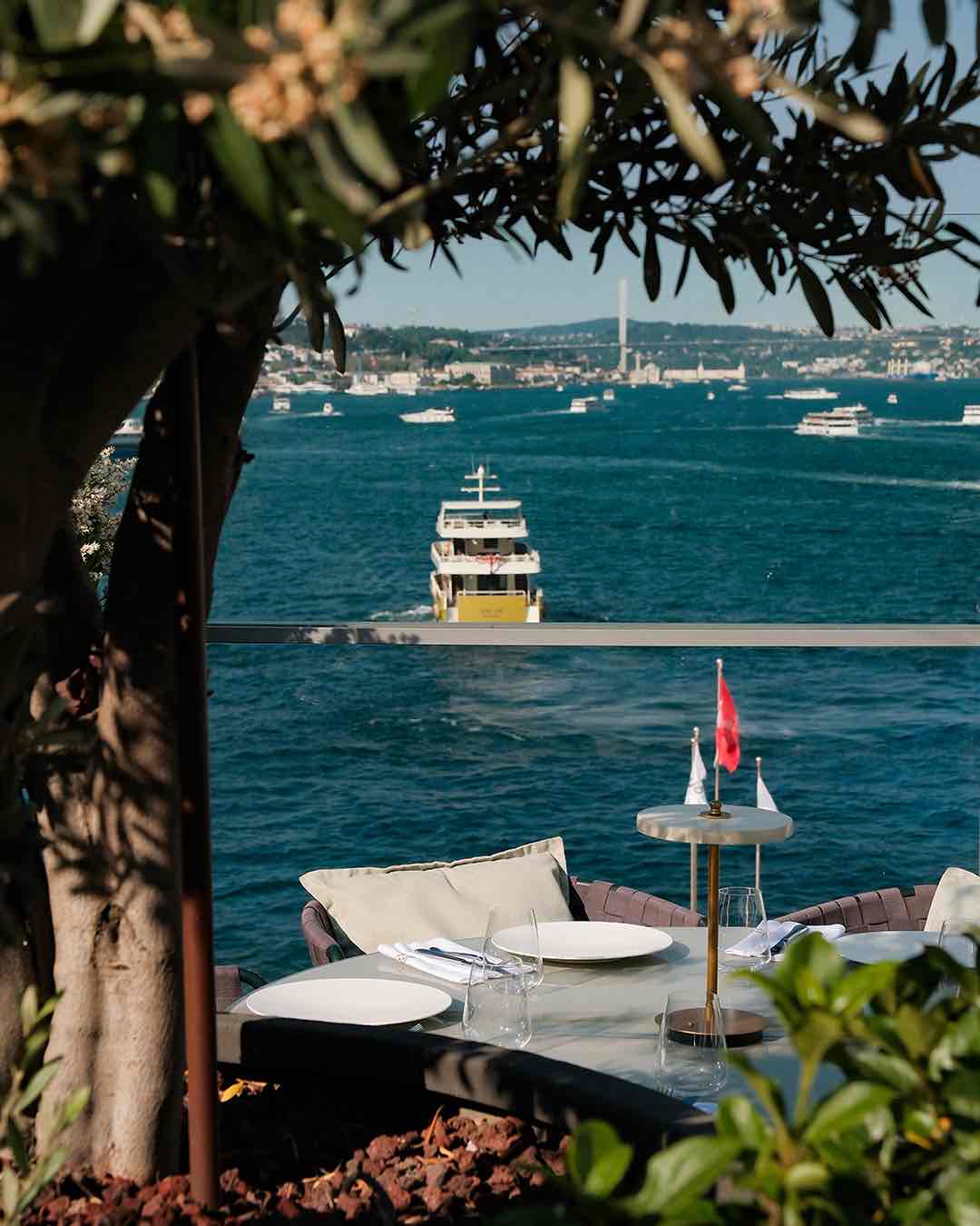 Ön planda yeşil yapraklarla çevrili, beyaz tabaklar, çatal bıçak takımları ve küçük bir Türk bayrağının yer aldığı şık bir masa; arka planda ise mavi denizde ilerleyen sarı-beyaz bir vapur ve İstanbul Boğazı'nın karşı kıyısındaki silüeti net bir şekilde görünüyor.