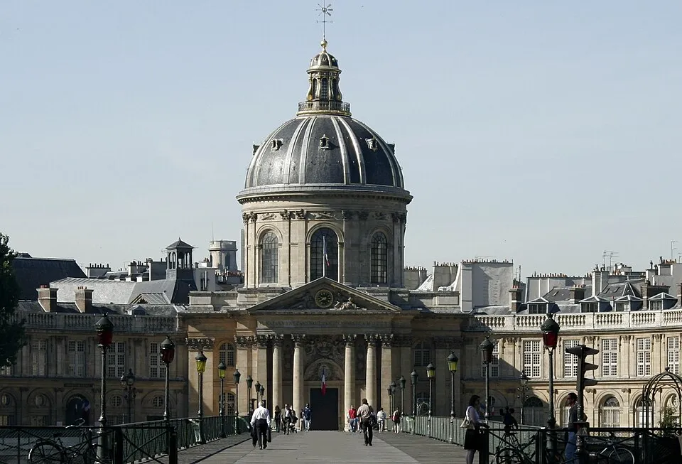 Paris'teki Pont des Arts köprüsünden bakıldığında, Fransız dil ve edebiyatında klasisizmin merkezi olan Académie Française'in büyük kubbesi, görkemli sütunları ve zarif detaylarıyla dikkat çeken klasik mimarisi ön planda, köprüde insanlar yürüyor.