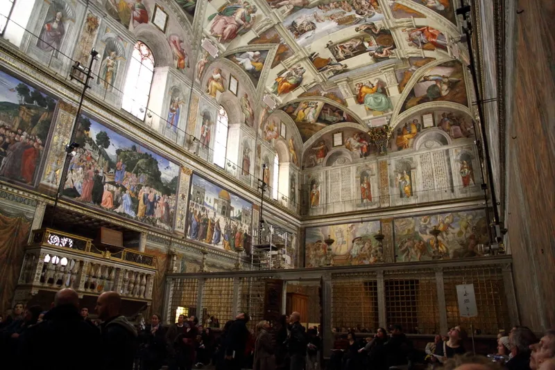 Vatikan'daki Sistine Şapeli'nin içinden çekilen bu geniş açılı karede, tavanı ve duvarları tamamen kaplayan, Michelangelo'nun başyapıtı olan göz alıcı dini freskler tüm ihtişamıyla sergilenirken, şapelin alt kısmında bu muazzam sanatsal güzelliği hayranlıkla izleyen kalabalık ziyaretçiler görülüyor.