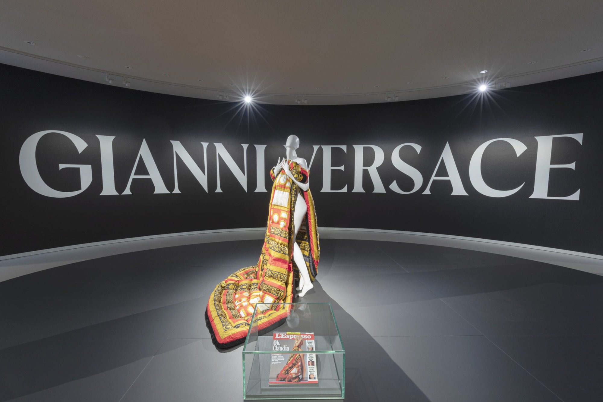 Gianni Versace sergisi kapsamında, büyük beyaz Gianni Versace yazısının yer aldığı siyah bir duvarın önünde, kırmızı, sarı, siyah ve altın tonlarında zengin desenlere sahip, yere kadar uzanan, kapitone ve gösterişli bir kostüm giymiş başsız bir manken ve önünde L'Espresso dergisinin sergilendiği cam bir vitrin yer alıyor.