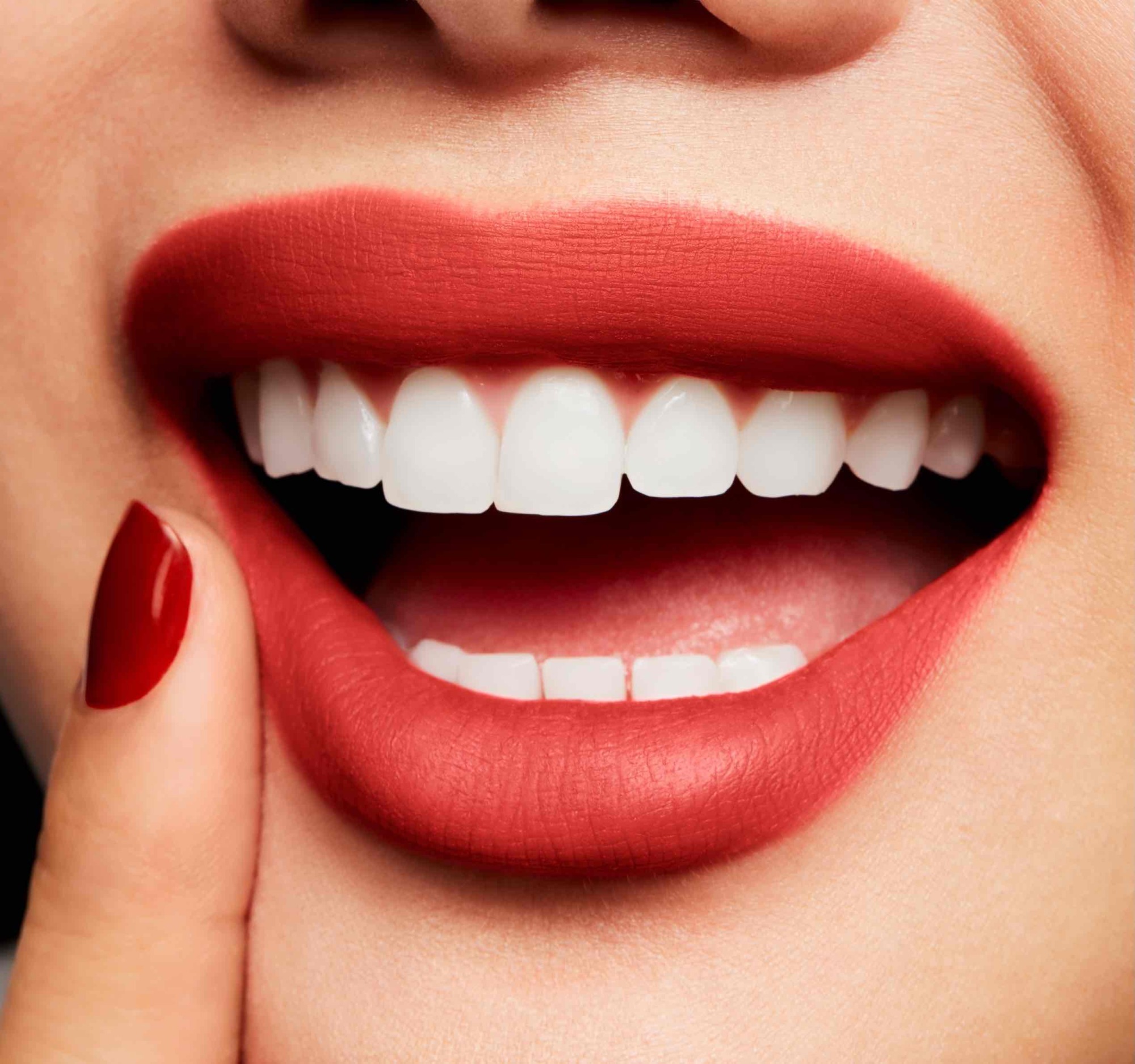 Puslu mat kırmızı ruj sürülmüş dudaklar ve beyaz dişlerle gülümseyen bir kadın. Kırmızı ojeli parmağıyla yanağına dokunarak rujun kadifemsi dokusunu ve canlı rengini öne çıkarıyor.