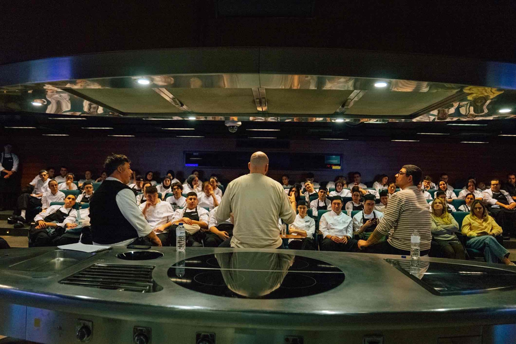 MSA'nın modern oditoryumunda, üç eğitmen şef, kendilerini dikkatle dinleyen aşçılık öğrencilerine bir seminer veriyor. Ön planda parlak, paslanmaz çelik bir gösteri mutfağı bulunuyor.
