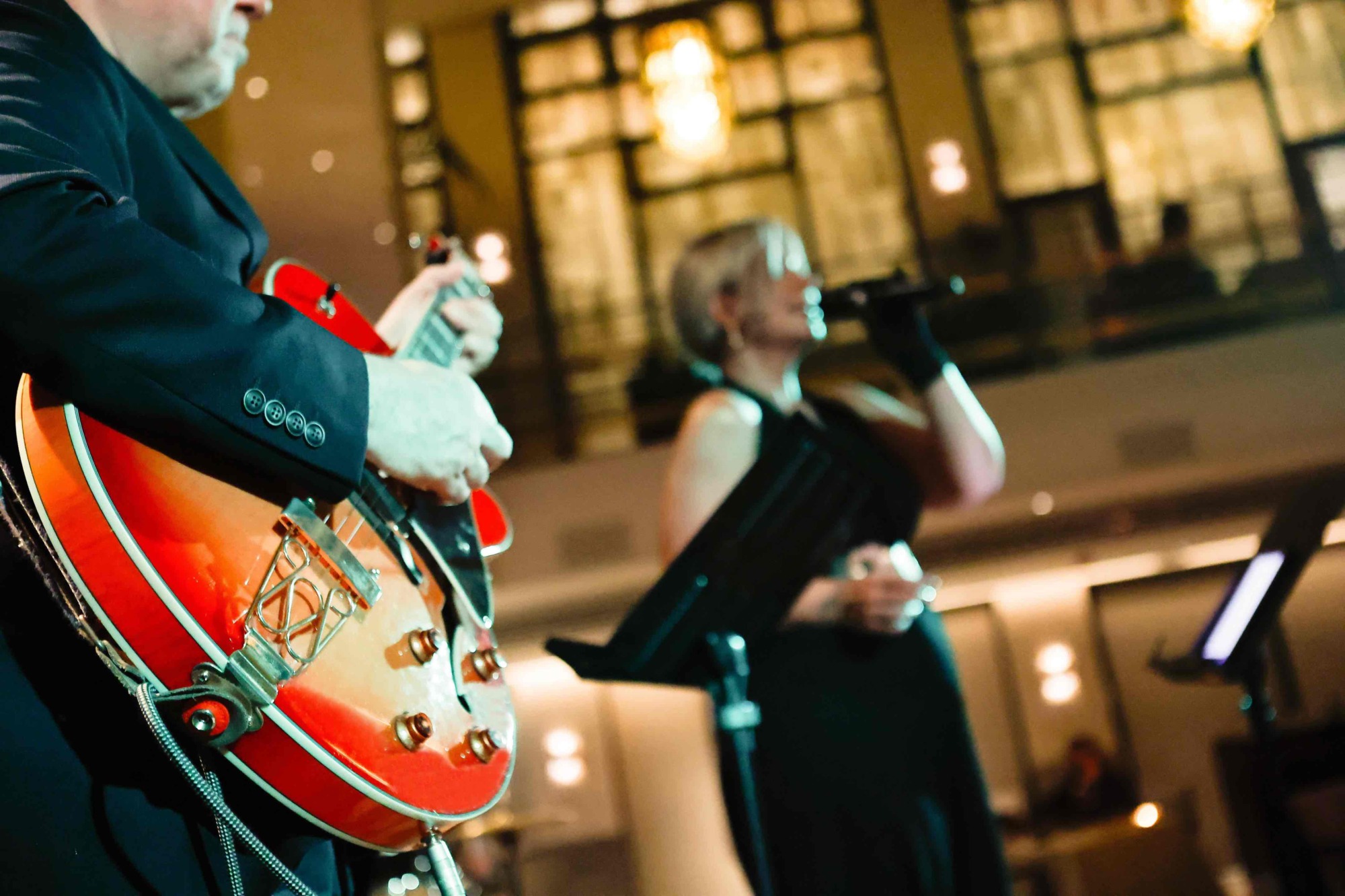 Ön planda, takım elbiseli bir müzisyen parlak kırmızı elektro gitarını çalarken, arka planda bulanık bir kadın vokalist mikrofona şarkı söylüyor; sıcak ışıklar sofistike bir atmosfer yaratıyor.