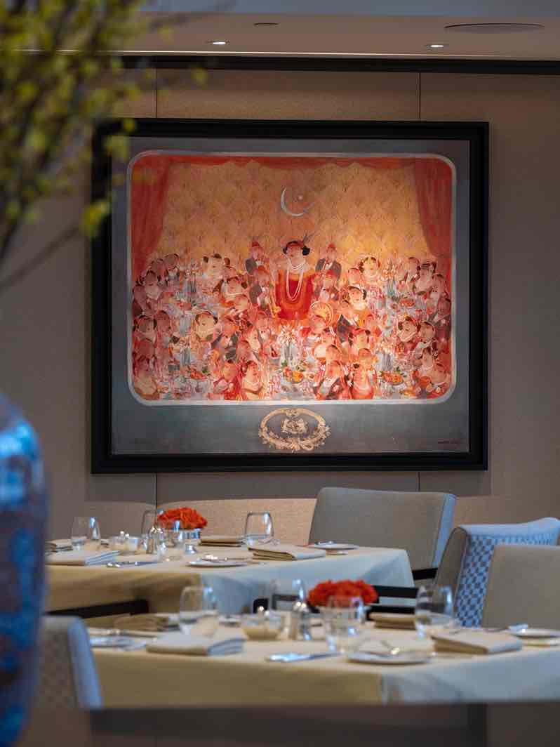 Geleneksel bir kalabalığı ve merkezi bir figürü tasvir eden büyük bir sanat eseri, şık bir restoranın duvarını süslüyor. Ön planda, beyaz örtülü ve çiçekli masalar zarif bir atmosfer yaratıyor.