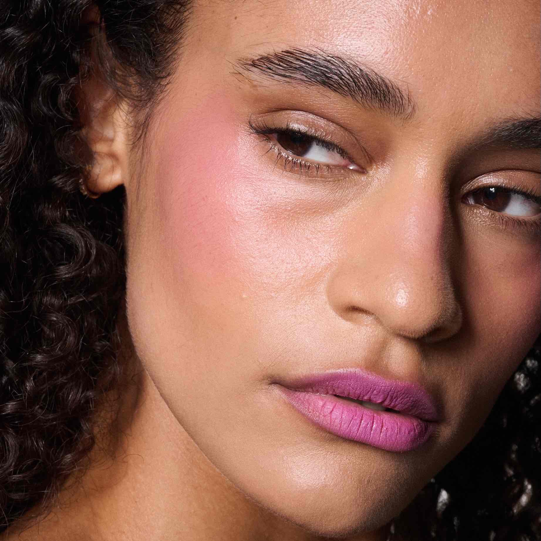 Esmer tenli model, dudaklarında Powder Kiss rujun kadifemsi dokusunu ve canlı pembe rengini sergiliyor. Yanaklarındaki allık ve ışıltılı cildiyle makyajı tamamlanmış.