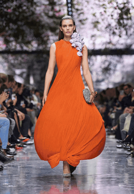 Haute couture defilesi podyum görünümü: Omuzda çiçek aplikeli, akışkan formda turuncu couture elbise ile sunulan dramatik tasarım.