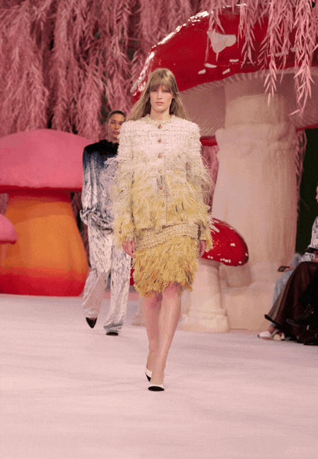 Chanel 2026 İlkbahar-Yaz Haute Couture defilesinde podyumdan bir görünüm: Masalsı mantar dekorları arasında sunulan, dokulu yüzeye ve püskül detaylara sahip sarı-beyaz couture elbise.